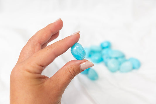 Hand holding a blue aragonite tumblestones with more crystals in the background on a white surface