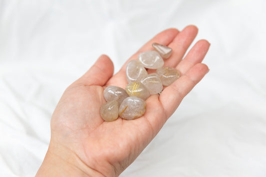 Hand holding small golden rutile quartz crystal tumeblstones against a white background
