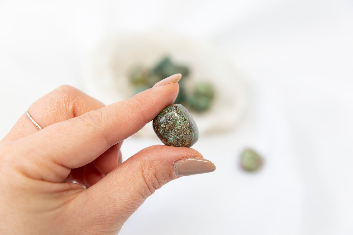 Hand holding a small green ruby fuchsite tumblestone against a blurred natural background